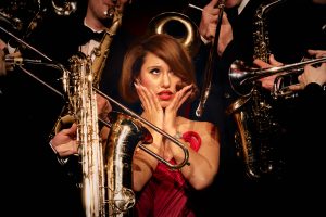 RAYE surrounded by brass instruments in red dress for This Music May Contain Hope press photo