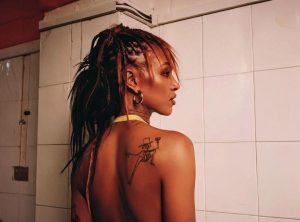 Alewya portrait with braided hair and back tattoo in tiled room, photographed by Bernice Mulenga