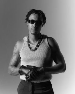 Skilla Baby poses in black and white wearing sunglasses, leather gloves, a chain necklace, and a ribbed tank top during a studio portrait session.
