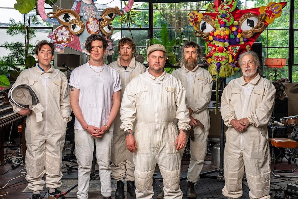 Modest Mouse members pose in white protective suits inside a colorful studio surrounded by oversized insect-inspired art installations