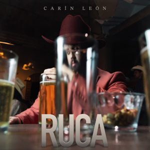 Carín León Ruca single cover showing artist in red suit reflected through glass at bar