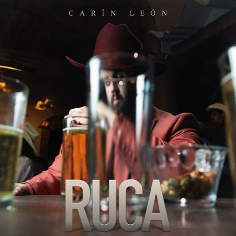Carín León Ruca single cover showing artist in red suit reflected through glass at bar