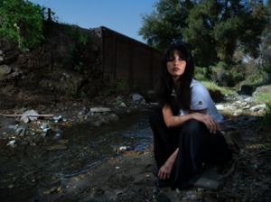Leyla Ebrahimi sitting by a creek in a wooded outdoor setting wearing a white tee and black pants