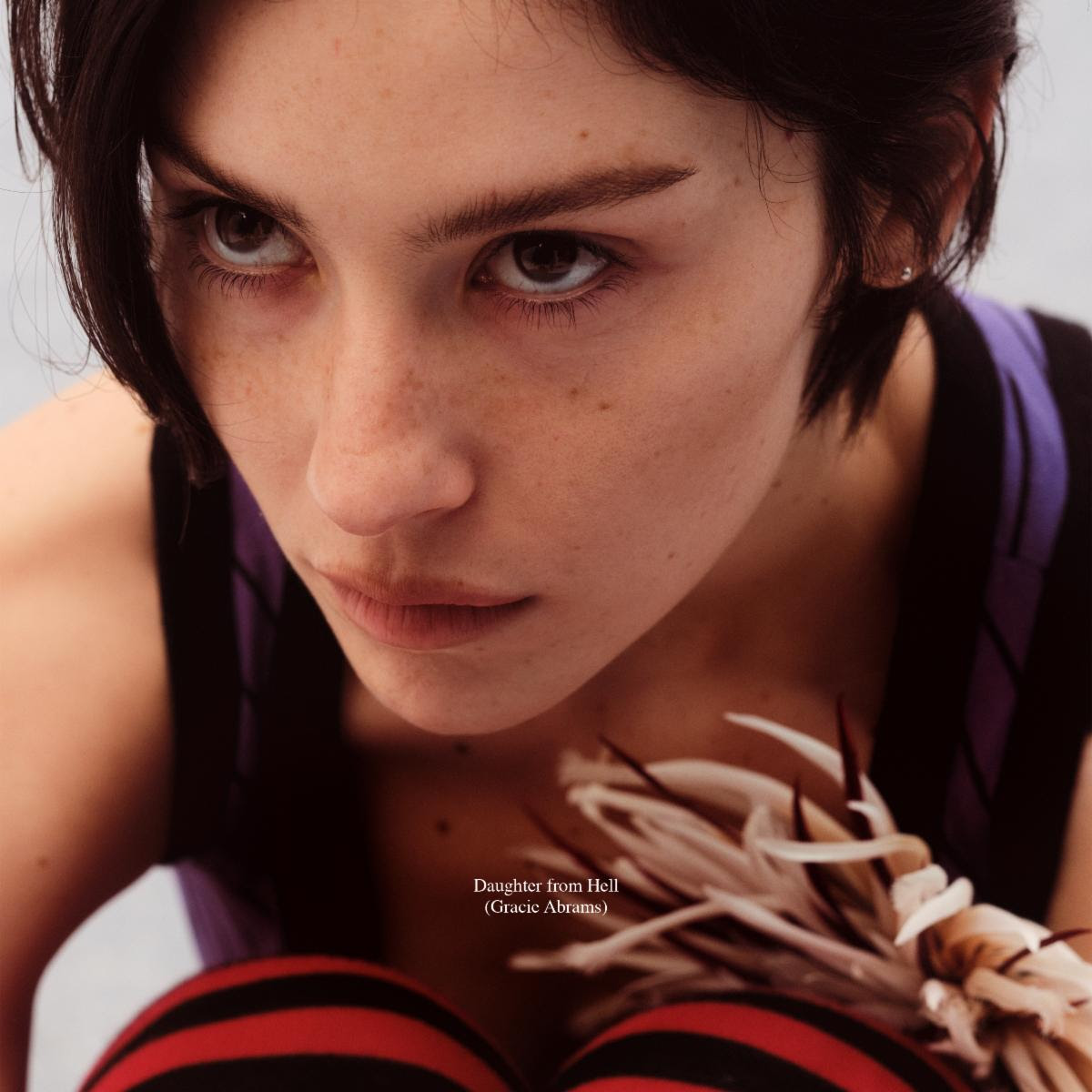 Gracie Abrams on the cover of Daughter from Hell wearing striped tights and holding flowers in a close-up portrait