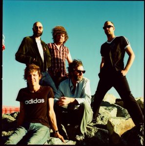 Members of Pond posing on coastal rocks under a bright blue sky in a retro-inspired band portrait