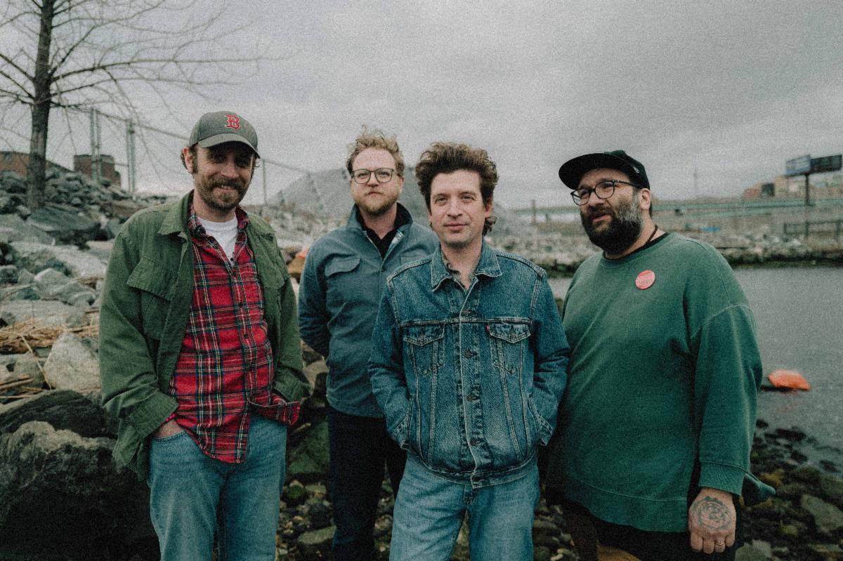 Members of Deer Tick posing outdoors near the water ahead of the release of their album ACI