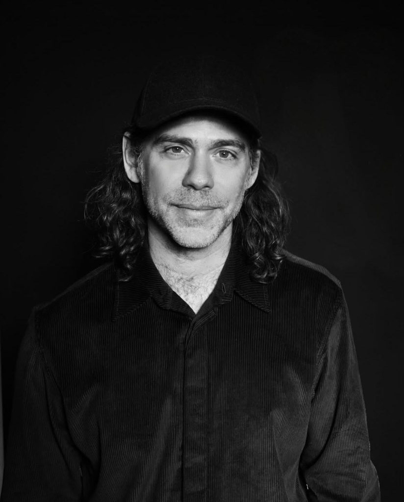 Producer Aaron Dessner in a black baseball cap and corduroy shirt photographed against a dark studio backdrop