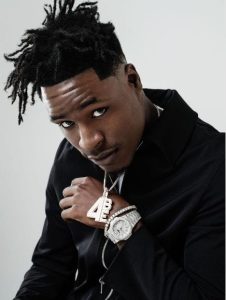 Mudbaby Ru poses in a black-and-white promotional portrait wearing a tank top, gloves, sunglasses, and chain necklace while kneeling against a studio backdrop.
