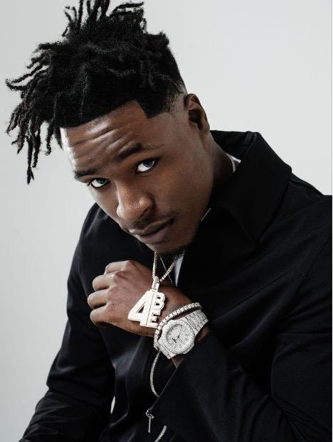 Mudbaby Ru poses in a black-and-white promotional portrait wearing a tank top, gloves, sunglasses, and chain necklace while kneeling against a studio backdrop.