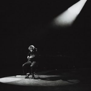 Lewis Capaldi sitting under spotlight performing solo on stage in black and white