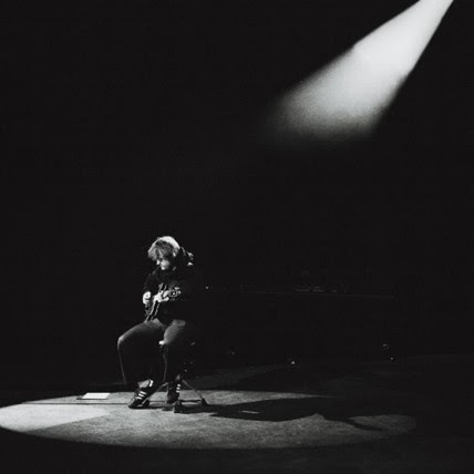 Lewis Capaldi sitting under spotlight performing solo on stage in black and white
