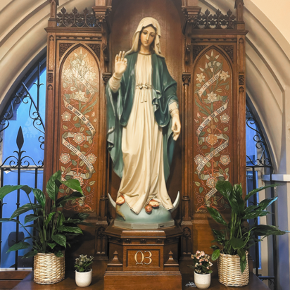 Virgin Mary statue displayed inside a church alcove with candles, plants, and ornate wood detailing