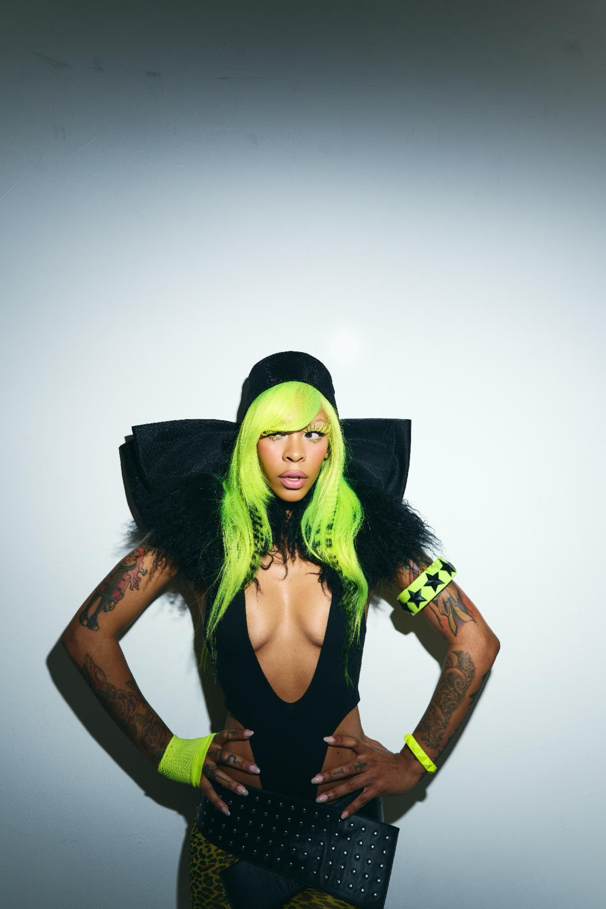 Rico Nasty poses in a black bodysuit with neon green hair and matching accessories against a white backdrop