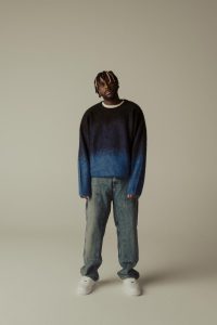 Myles Smith standing in a blue gradient sweater and jeans during a promotional photo shoot for his debut album My Mess, My Heart, My Life.