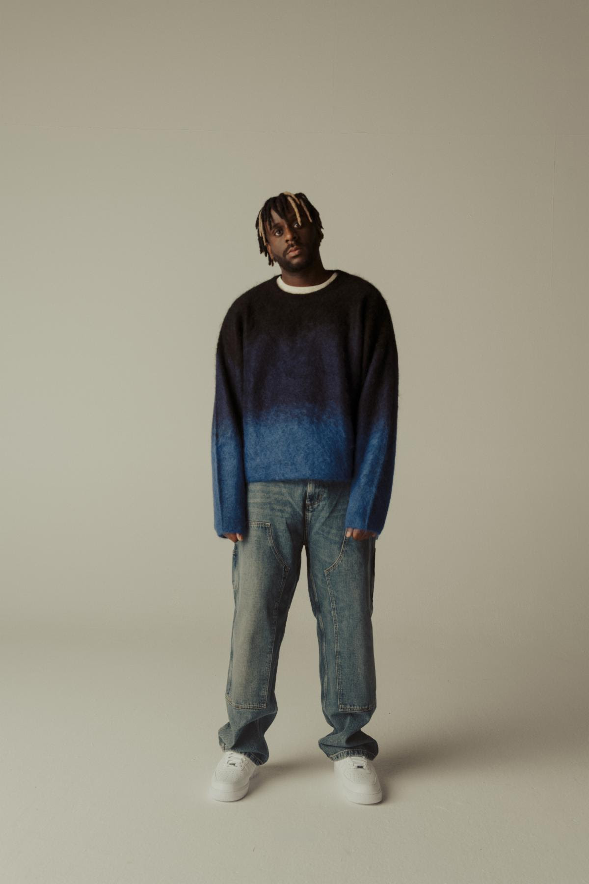 Myles Smith standing in a blue gradient sweater and jeans during a promotional photo shoot for his debut album My Mess, My Heart, My Life.