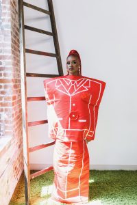 Tierra Whack poses in a sculptural red outfit inspired by a suit design beside a wooden ladder during the WHACK'S MUSEUM promotional campaign