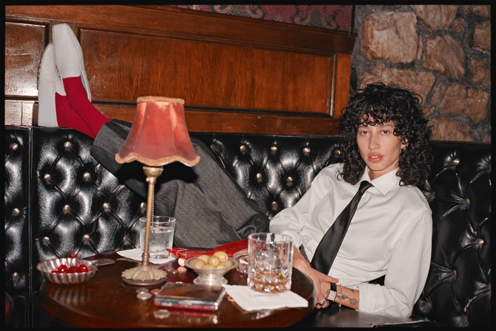 Towa Bird lounges in a restaurant booth wearing a white shirt and black tie in a promotional portrait for Gentleman