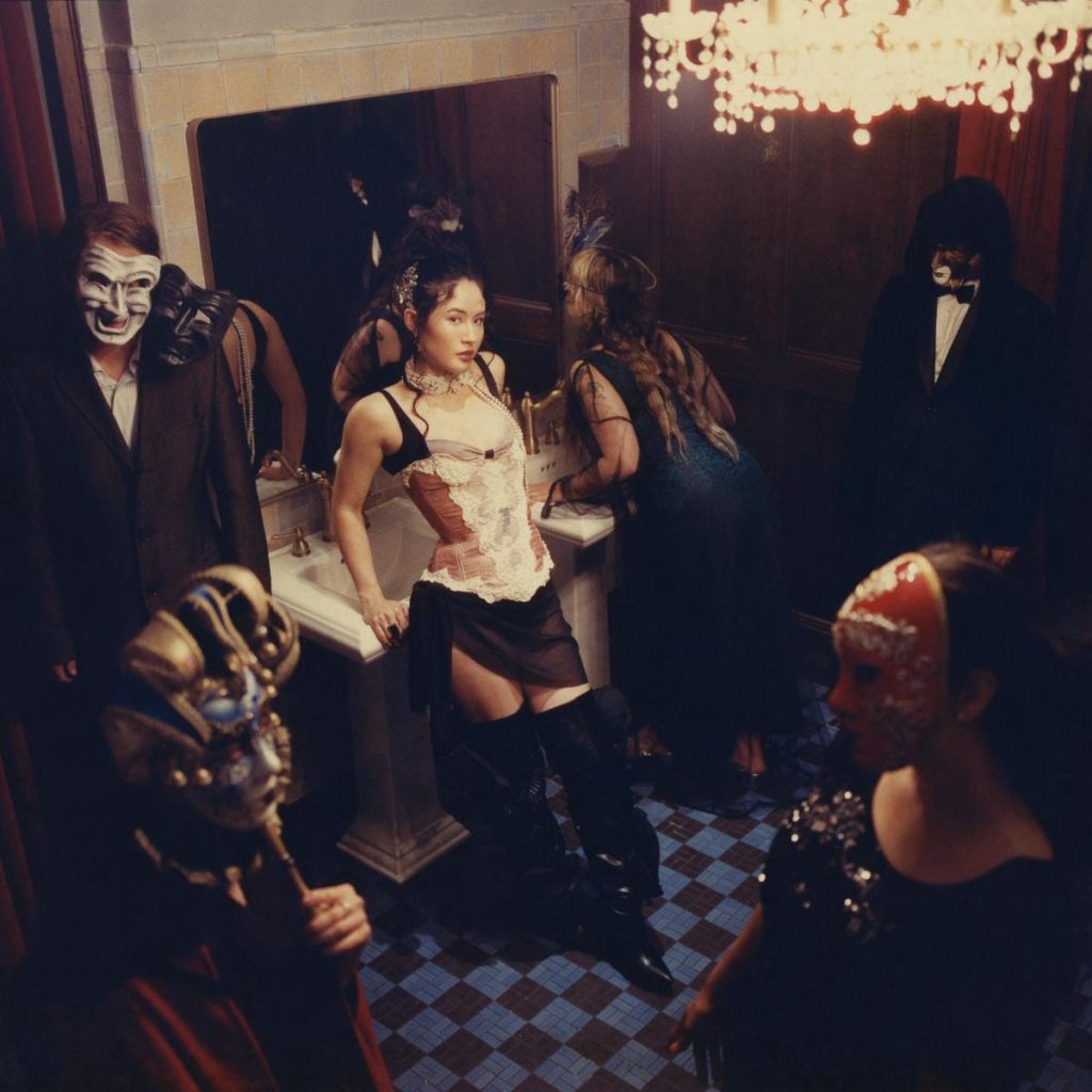 Emei poses in a vintage corset and black boots inside a dramatic masquerade-inspired ballroom surrounded by masked figures for her Night at the Opera EP campaign.