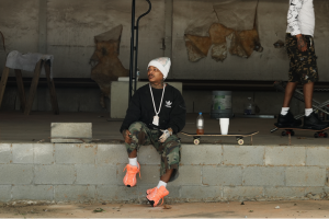 Rylo Rodriguez sits beside a skateboard in an industrial setting ahead of the release of his album S.K.A.T.E. (Silence Keeps All Targets Exposed).