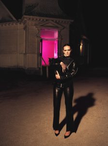 Tove Lo stands in black leather holding a black cat outside a building illuminated by pink light in a promotional photo for her 2026 album ESTRUS.