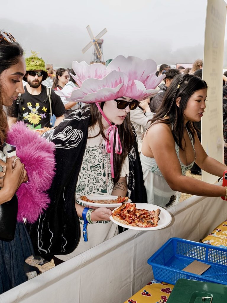 Festivalgoer carries two slices of pizza while wearing a giant pink flower hat at Outside Lands 2026 in San Francisco.