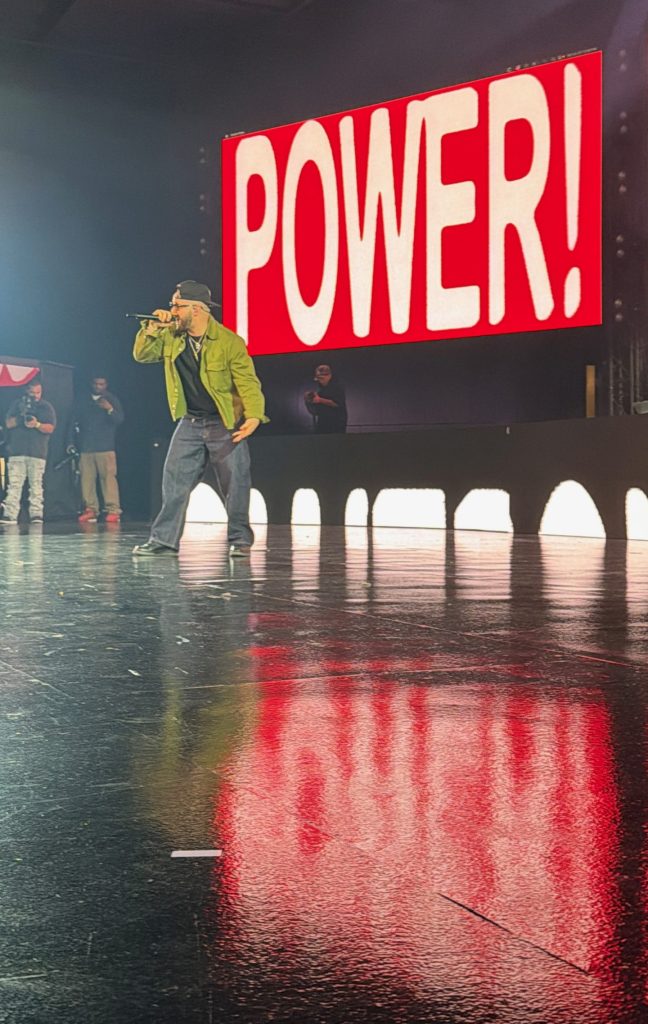 Andy Mineo performs at the edge of the stage while fans raise their phones and hands.