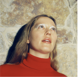 Julia Jacklin wearing a red turtleneck and looking upward against a stone wall.