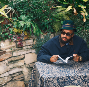 Larry June reading at an outdoor table in a promotional photo for his album Who Coppin?