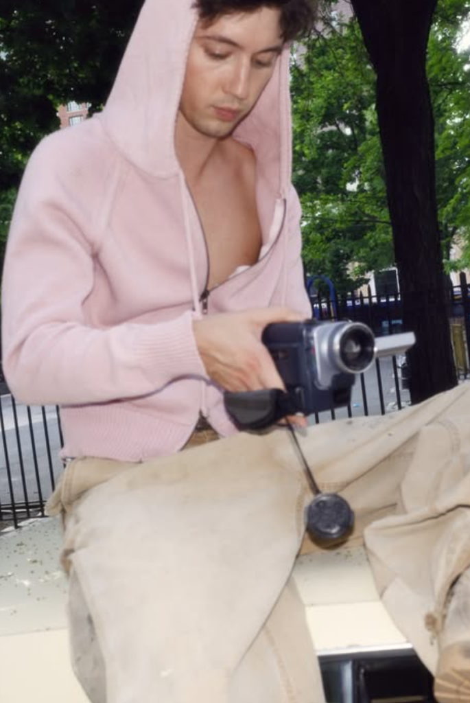 Troye Sivan holding a handheld camcorder outdoors while wearing a pale pink hoodie.