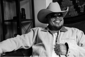 Maoli smiles while wearing a cowboy hat, sunglasses, and a light denim jacket in a black-and-white promotional portrait ahead of the release of MMO3.