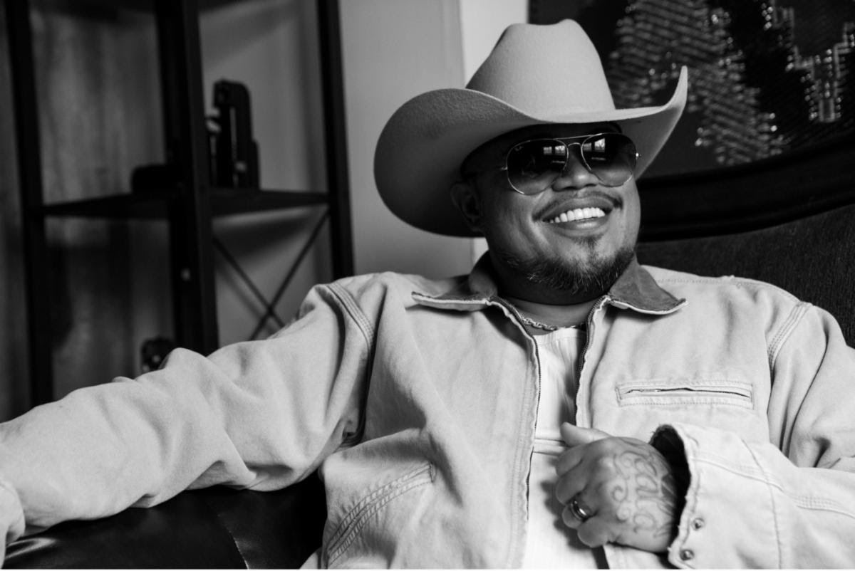 Maoli smiles while wearing a cowboy hat, sunglasses, and a light denim jacket in a black-and-white promotional portrait ahead of the release of MMO3.