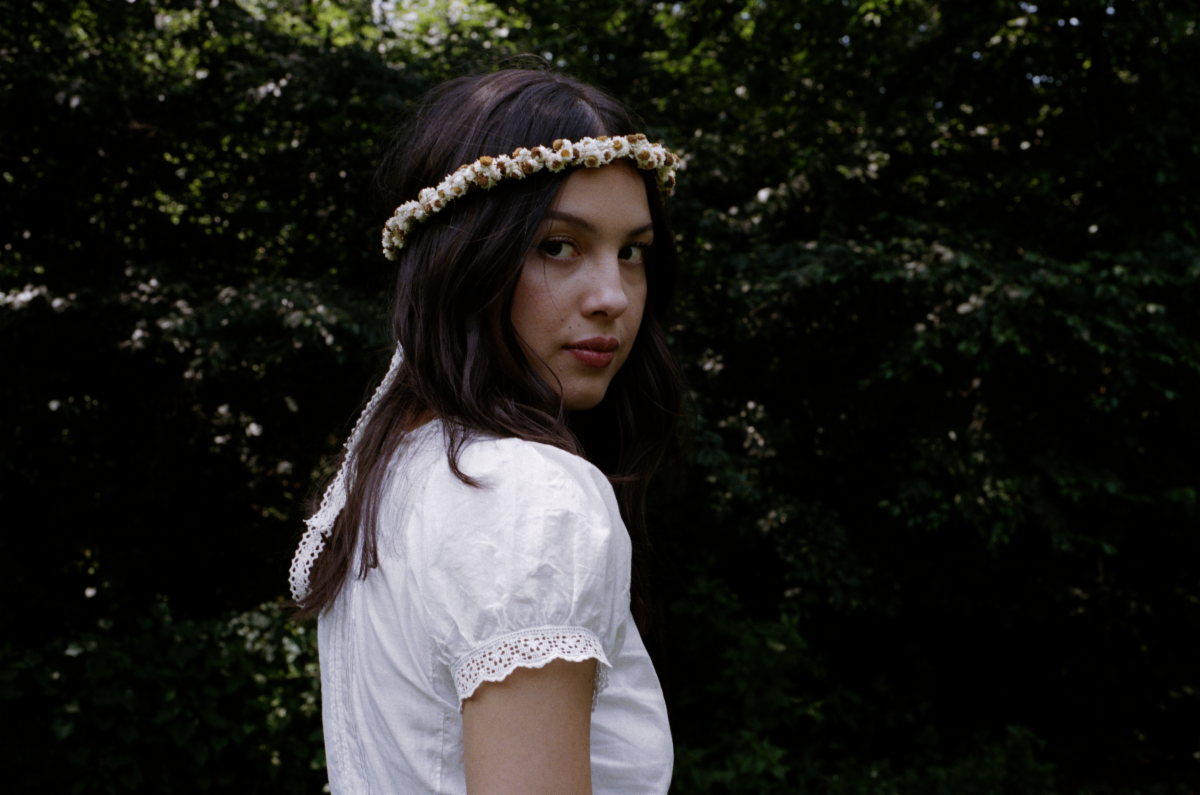 Olivia Rodrigo wearing a white dress and flower crown outdoors in a wooded setting.