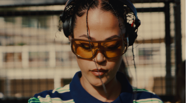 Irish dance artist Jazzy wearing headphones and amber-tinted sunglasses, looking downward in a close-up from her Dublin to the World documentary.