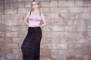 Blondshell singer Sabrina Teitelbaum wearing a pink satin top and black skirt against a stone wall for her 2026 album Violins