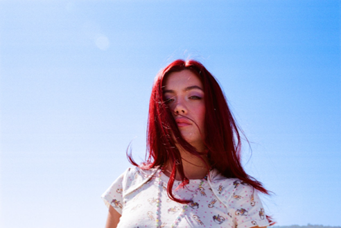 Alessi Rose with long red hair and a white floral-patterned top photographed from a low angle against a bright blue sky.