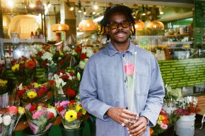 Pauli The PSM wearing glasses and a blue jacket, holding a pink rose while standing in front of colorful flower bouquets.