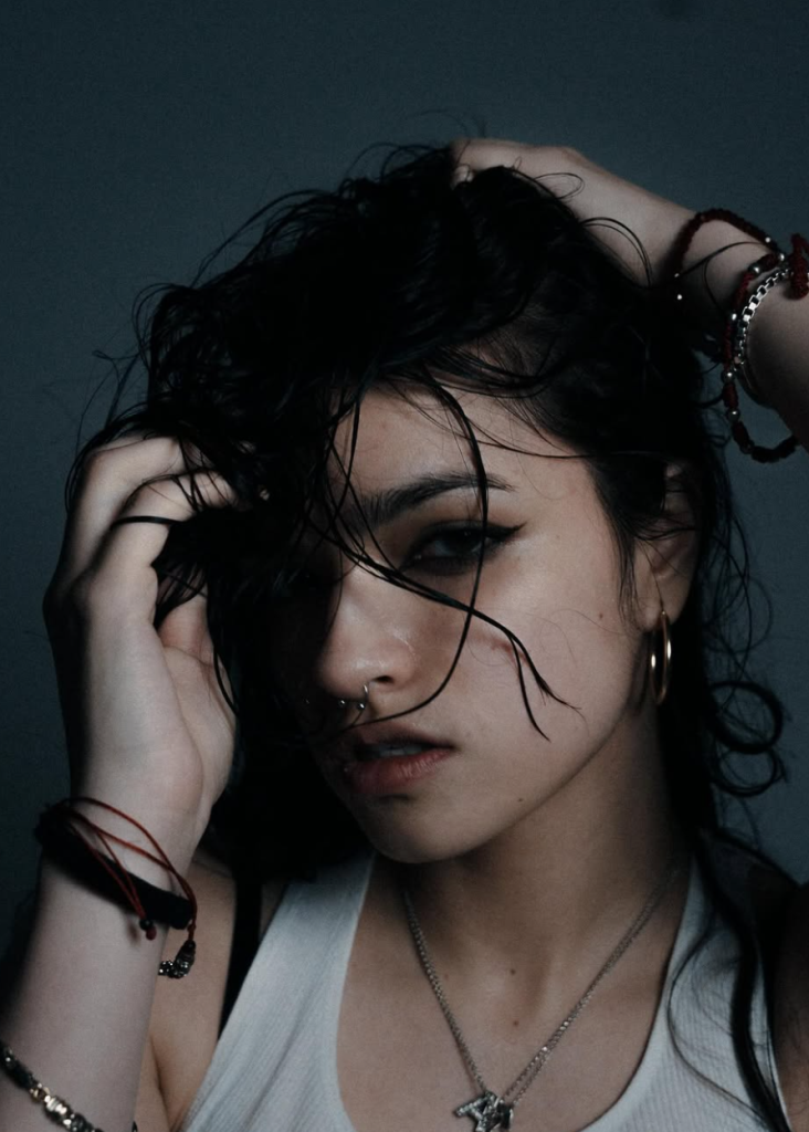 Ari Abdul with dark tousled hair, dramatic winged eyeliner, a nose piercing, and silver jewelry against a dark gray background.