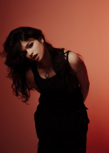 Ari Abdul wearing a black sleeveless outfit with long dark hair against a warm red-orange background.