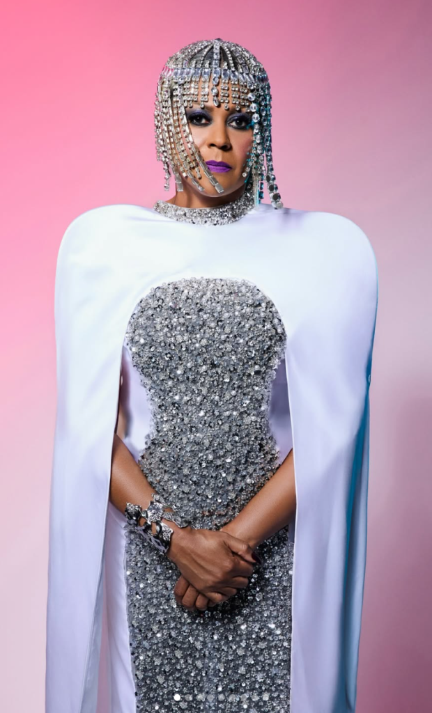Crystal Waters in a silver embellished gown, jeweled headpiece and white cape against a pink background.