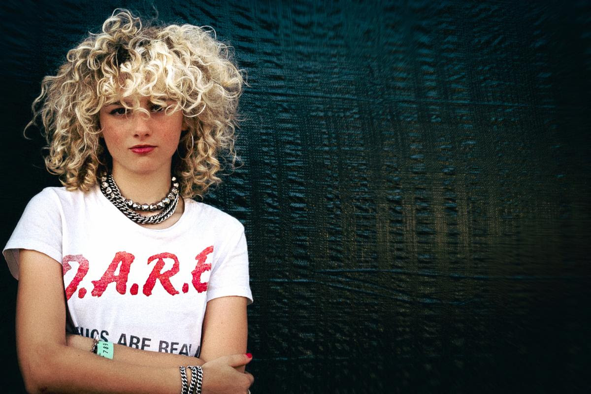 Guitarist and singer Grace Bowers with voluminous curly blonde hair, wearing a white D.A.R.E. T-shirt and silver chain necklaces against a dark background.