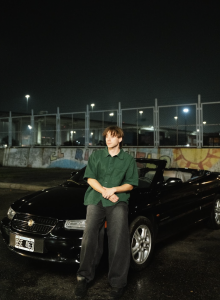 Paulo Londra wearing a green shirt and leaning against a black convertible at night, with graffiti-covered walls in the background.