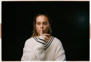 Tash Sultana wearing a white knit sweater against a black background, holding a finger to their lips.