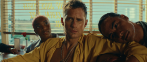 Alexander Skarsgård wearing a mustard-yellow shirt sits between two women in a diner scene from the Rolling Stones' “Mr Charm” music video.