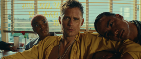 Alexander Skarsgård wearing a mustard-yellow shirt sits between two women in a diner scene from the Rolling Stones' “Mr Charm” music video.