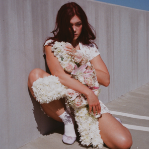 Alessi Rose crouches against a concrete wall wearing a white floral outfit and holding an arrangement of white chrysanthemums and pale pink roses.