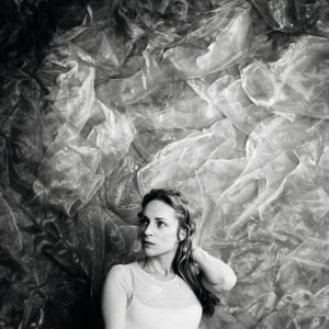 Black-and-white promotional portrait of Agnes Obel looking to the side with one hand resting in her hair against a textured floral backdrop.