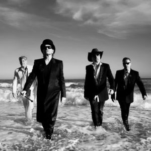 U2 band members walking through ocean surf in black and white, wearing suits, hats, and sunglasses