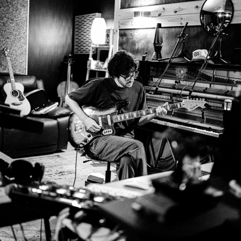 Australian producer and multi-instrumentalist Ethan French playing an electric guitar in a recording studio beside an upright piano, surrounded by instruments and recording equipment.