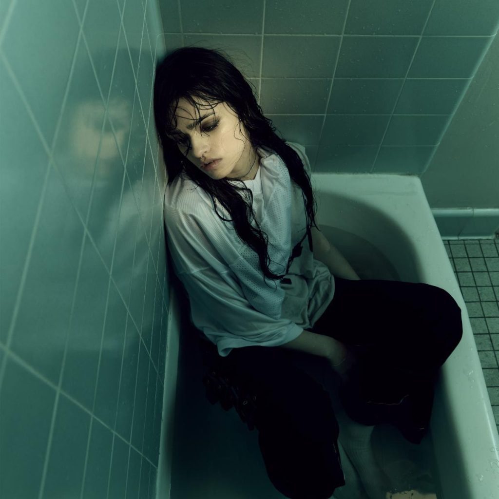 Ari Abdul sitting fully clothed in a white bathtub with wet dark hair, surrounded by green-tinted bathroom tiles.